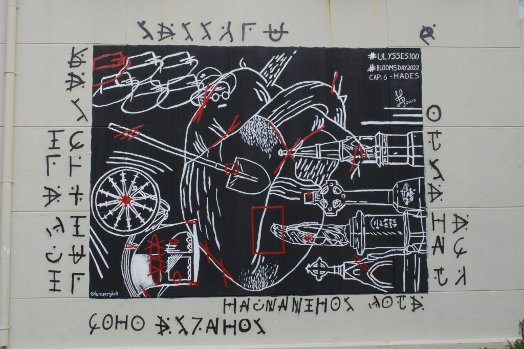 Photo description: The picture shows a wall with a grafitti on it. The grafitti is a black square with some white draws in it. It is possible to identify a human heart, a shovel, some hats, a compass, grave patters such as the celtic cross, a saint, and a bell tower. Around the picture there are a few symbols.