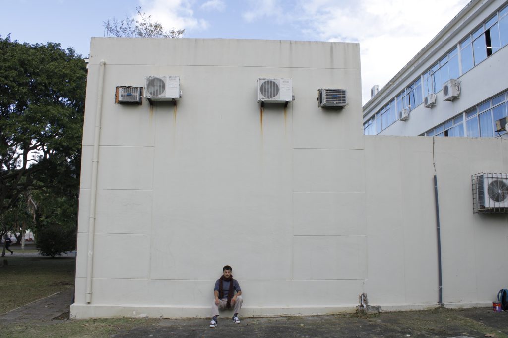 Photo showing a man with short dark hair sitting near the outside wall of a building. He wears a gray t-shirt, beige pants and a red scarf.