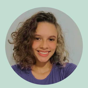 Photo of Julia, she has short curly hair, with half of it tied blond and half of it naturally burnet, She also has dark eyes. She is smiling at the camera and wearing a violet shirt.
